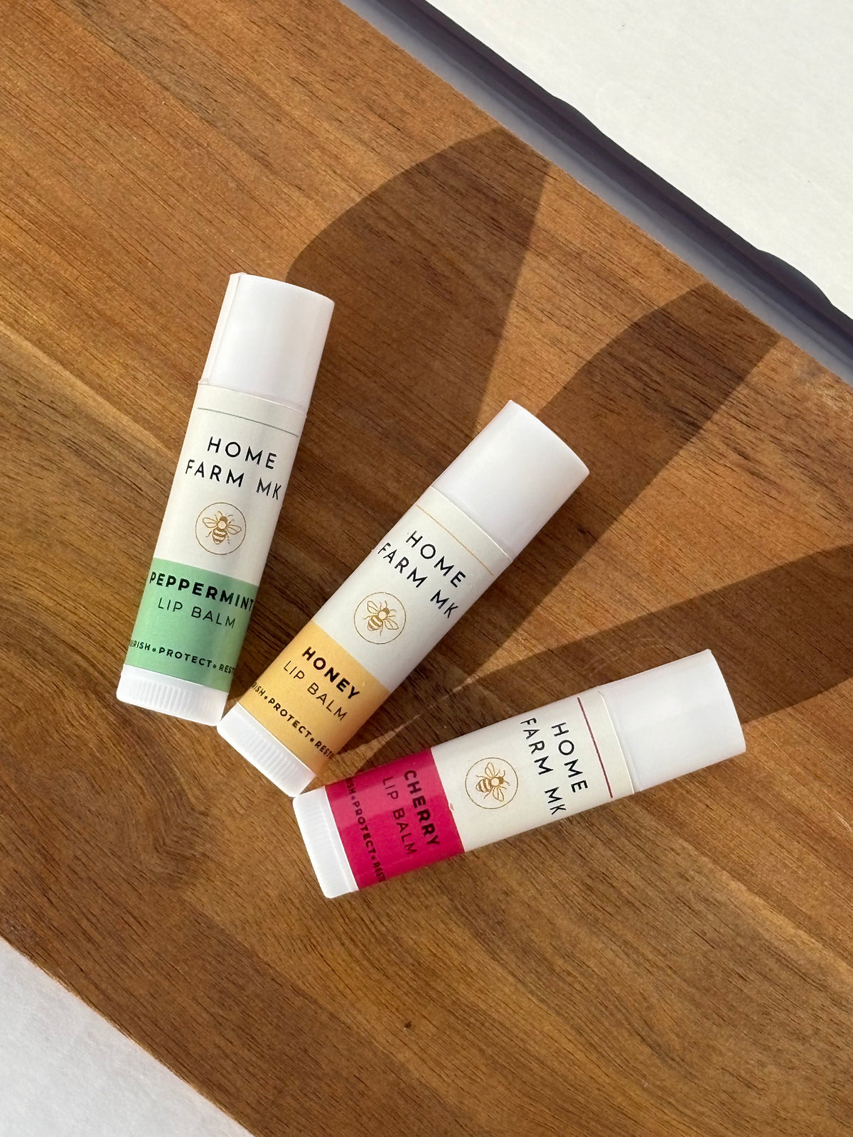 Home Farm MK Peppermint, Honey and Cherry lip balm tubes on a wooden surface.