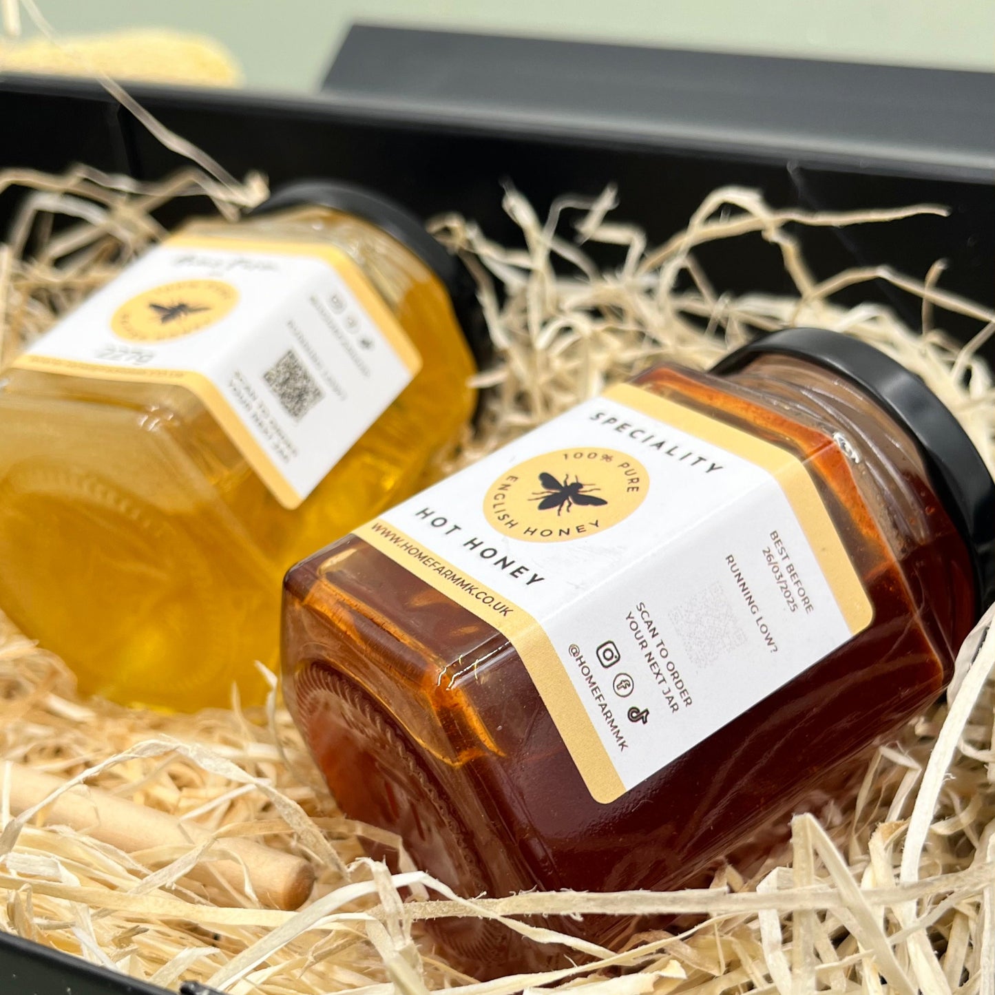 Two jars of flavoured honey on a bed of wooden straw in a black gift box