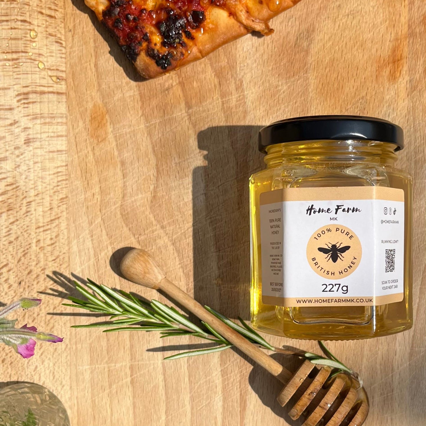 Jar of honey with a honey dipper on a wooden surface next to a slice of hot spicy pizza.