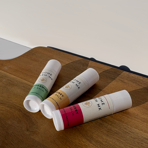 Three Beeswax Lip Balm tubes on a wooden surface. One has a white and green label for ‘Peppermint’ flavour, the second has a white and yellow label for ‘Honey’ flavour, and the third has a dark pink and white label for ‘Cherry’ flavour