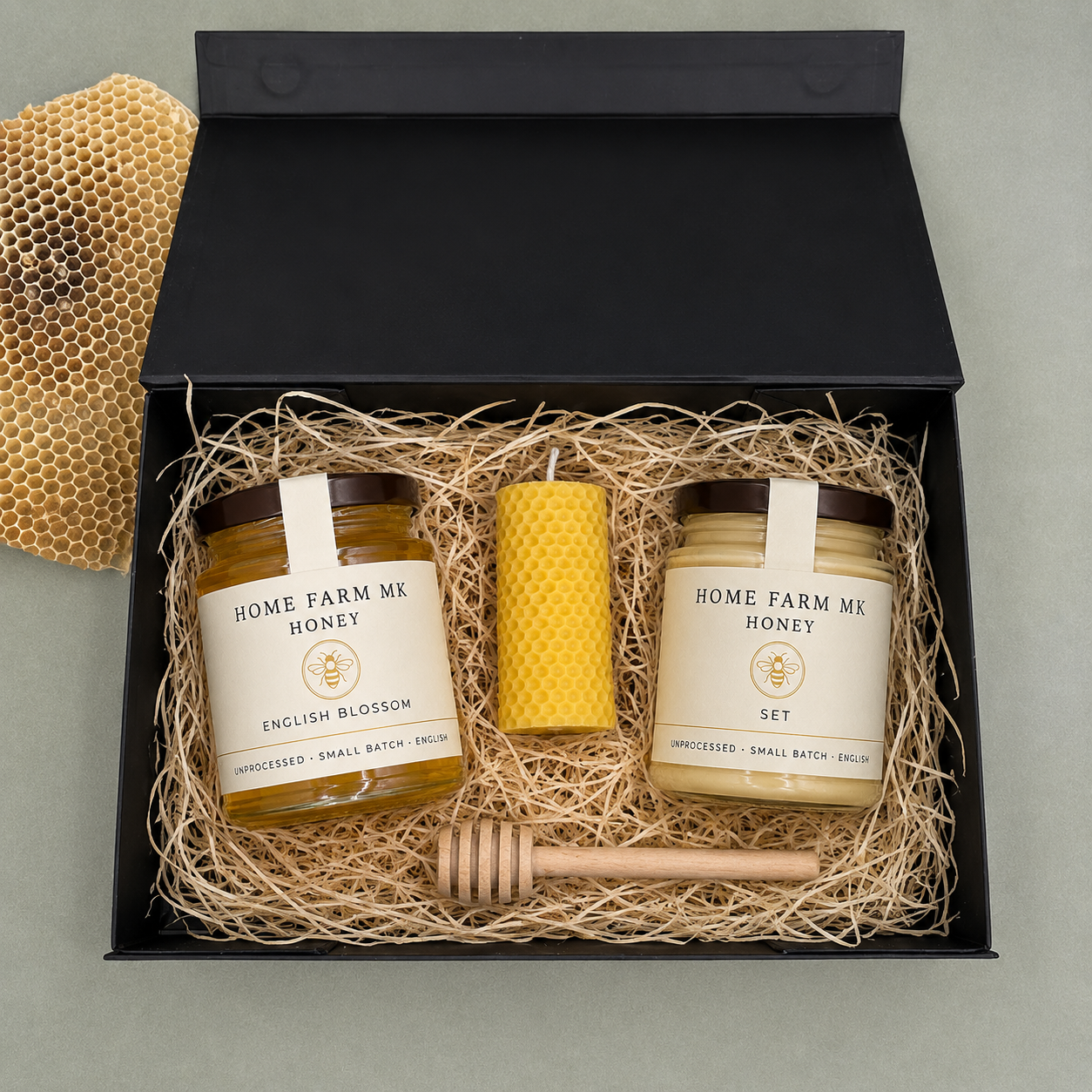 A gift set in a black box filled with shredded paper, containing two jars of 'Home Farm Mix' English Blossom honey, a yellow honeycomb candle, a wooden honey dipper, and a honeycomb prop on a gray background.