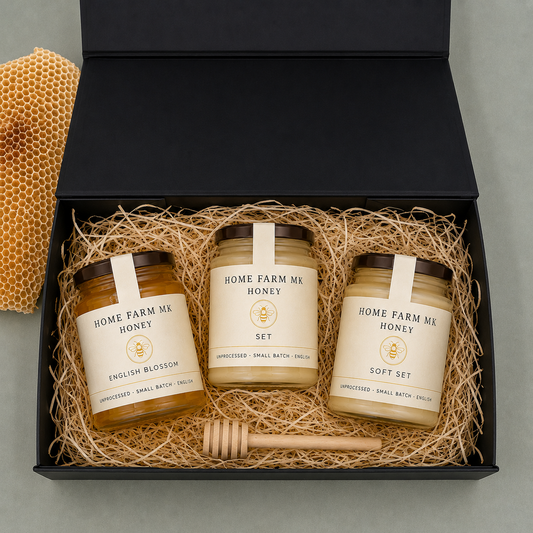 A gift set of three jars of honey from Home Farm Mk set in a black presentation box with shredded paper. The jars are labeled English Blossom, Soft Set, and White Set, with a wooden honey dipper and a piece of honeycomb visible.