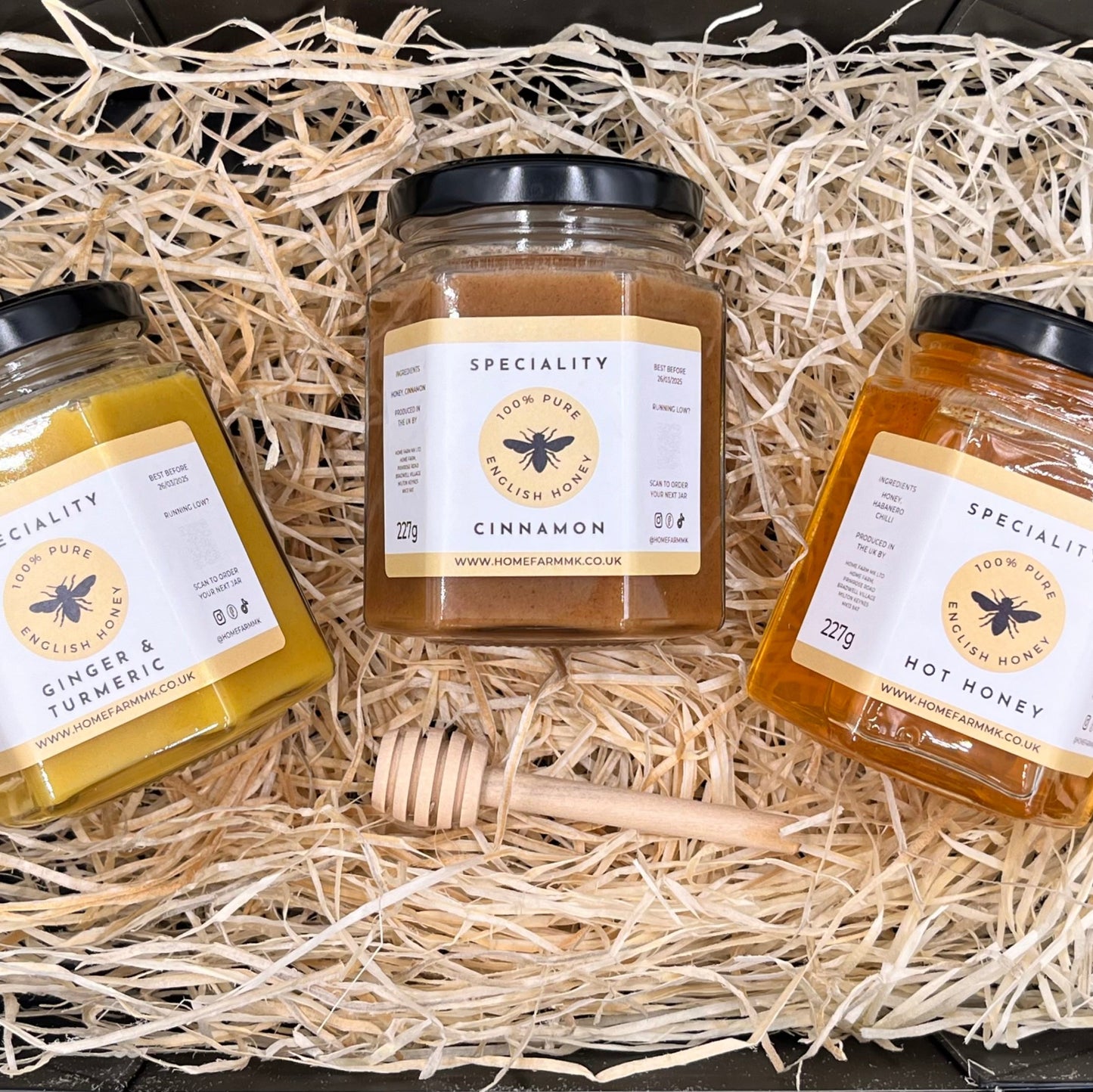 A jar of yellow ginger & turmeric honey, a jar of brown cinnamon honey, and a jar of hot habanero honey in a black gift-box, accompanied by a wooden honey dipper.
