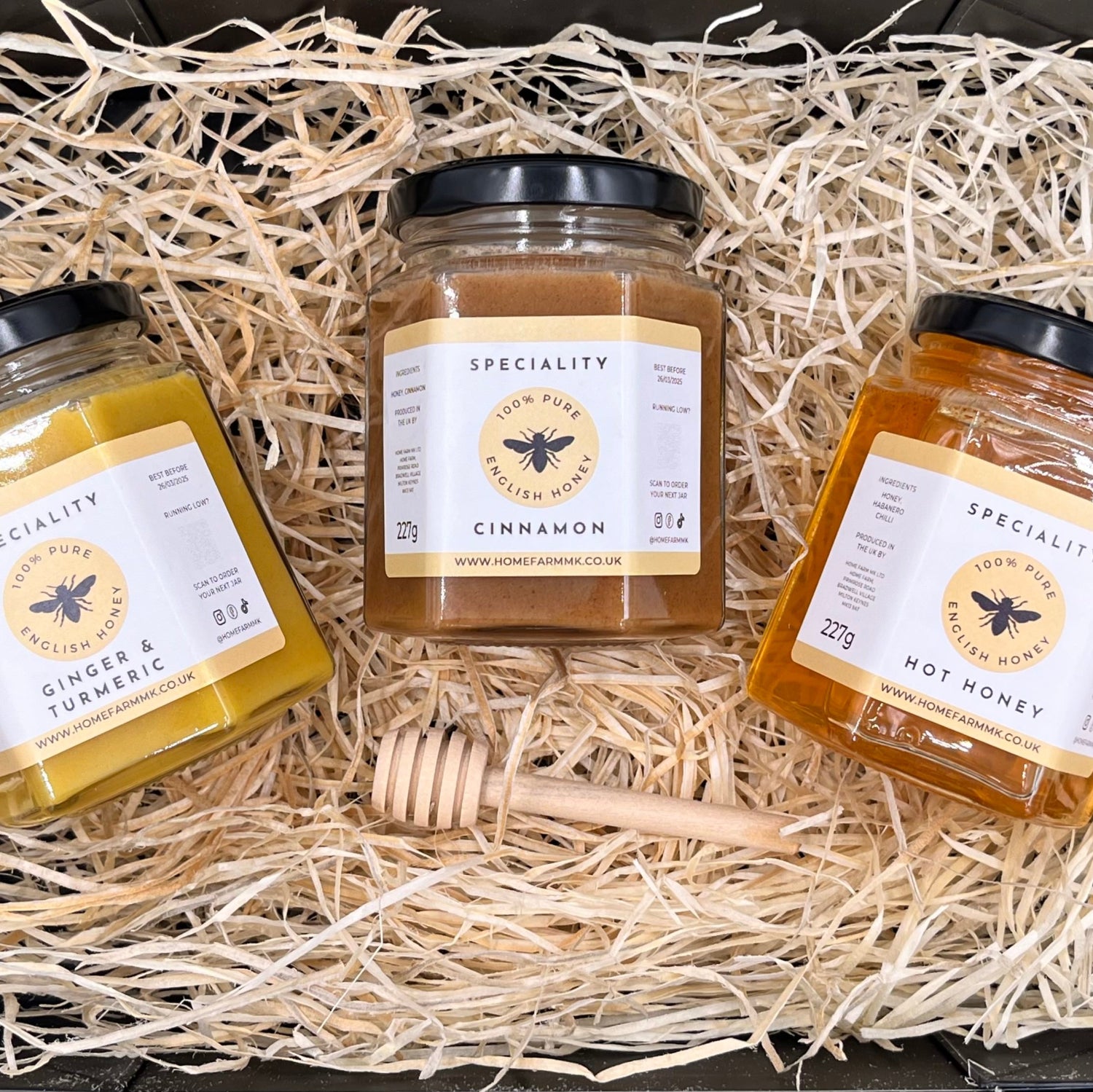 A jar of yellow ginger & turmeric honey, a jar of brown cinnamon honey, and a jar of hot habanero honey in a black gift-box, accompanied by a wooden honey dipper.