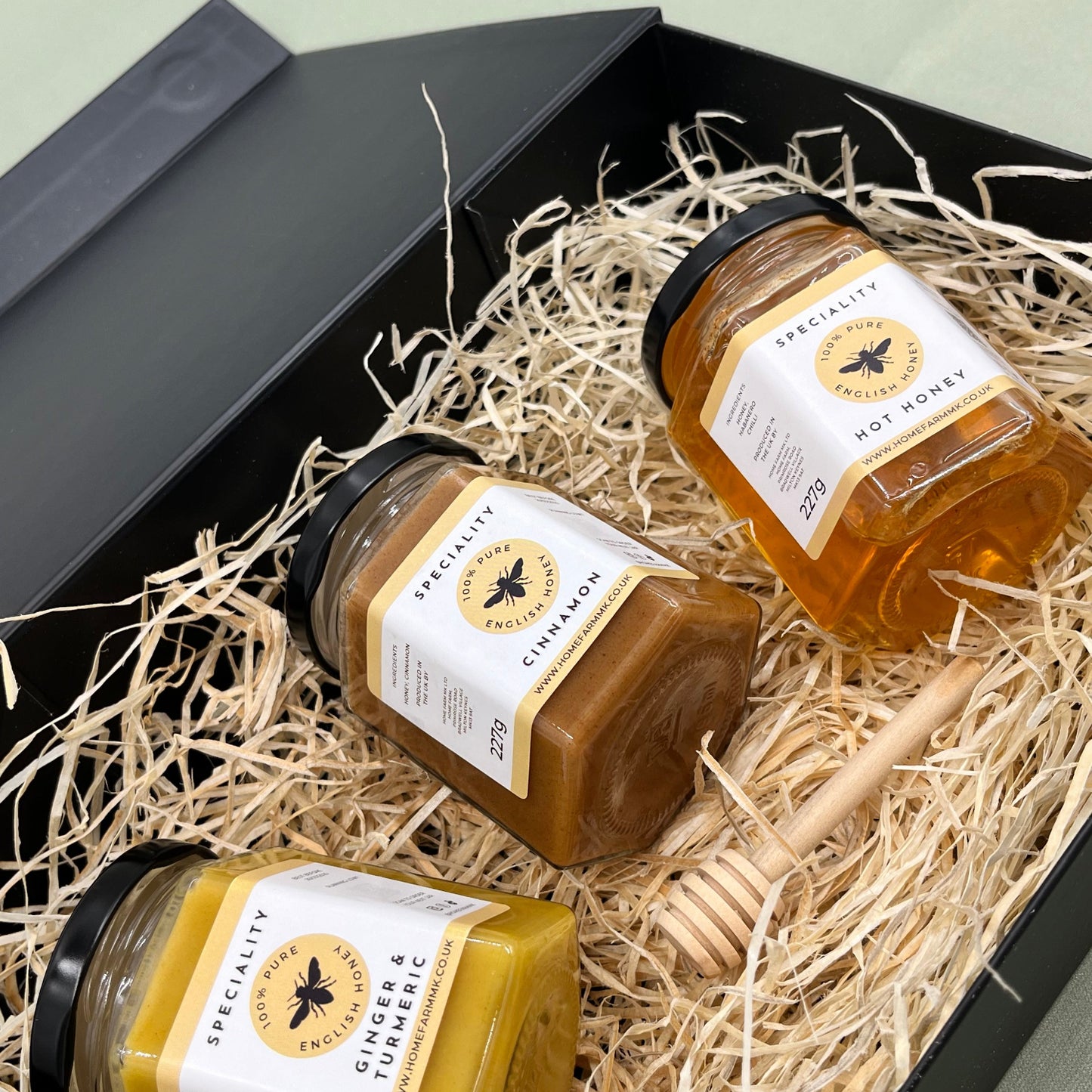A jar of yellow ginger & turmeric honey, a jar of brown cinnamon honey and a jar of orange hot habanero honey presented on a bed of straw in a black gift-box.
