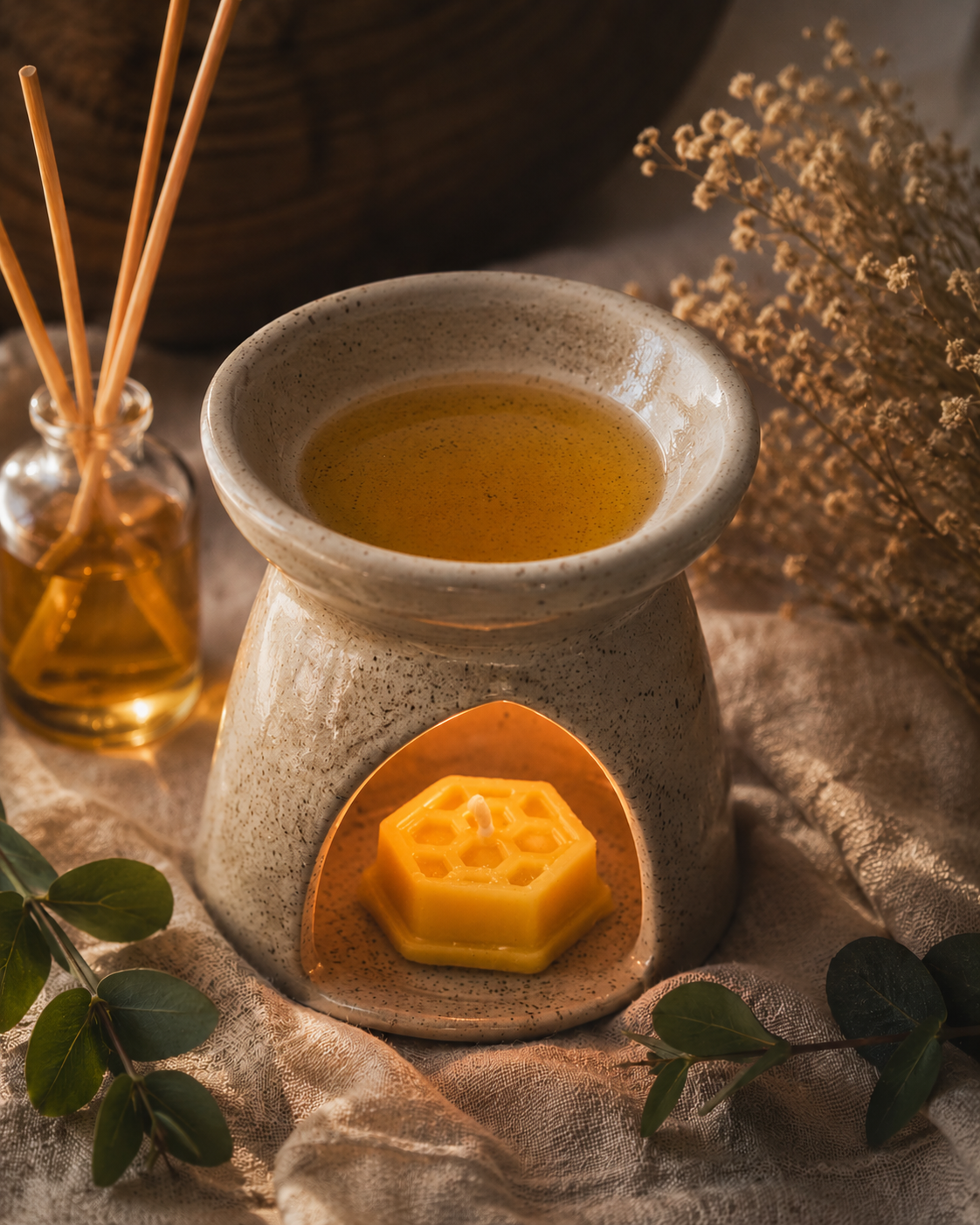 A ceramic wax warmer with a small yellow hexagonal wax melt inside, styled on textured beige fabric with dried flowers, green leaves, and a glass reed diffuser in the background.