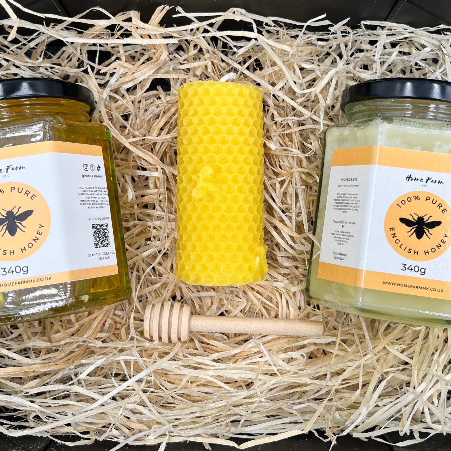 Two jars of honey, a yellow honeycomb pillar candle, and a honey dipper on a bed of wood straw.