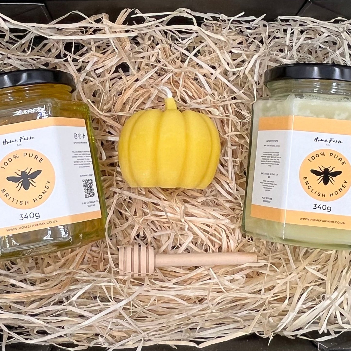 Two jars of honey with a small yellow pumpkin candle and a wooden dipper in a black gift box