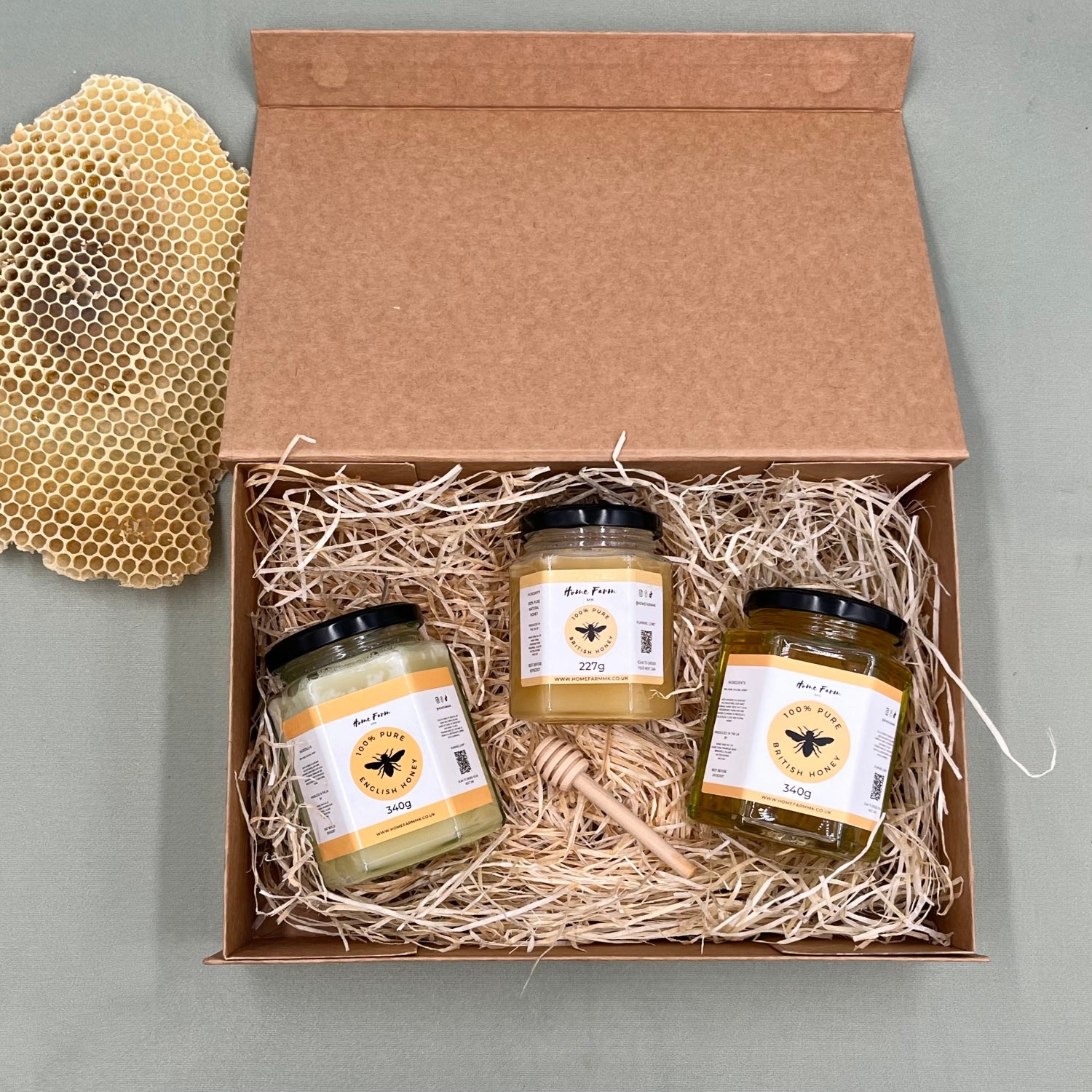 Three jars of honey and a wooden honey dipper in a brown kraft gift box, with honeycomb