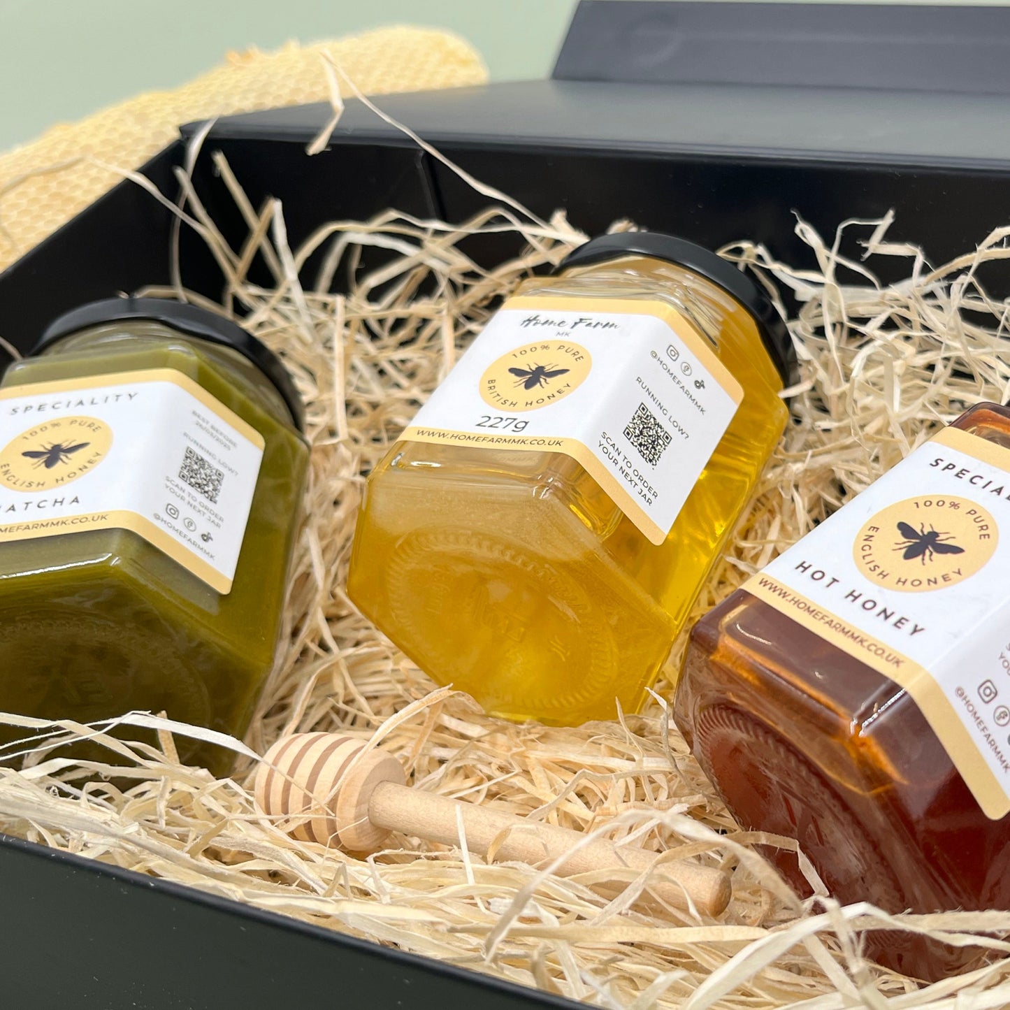 A jar of green matcha honey, a jar of yellow runny English honey and a jar of red hot honey, with a wooden honey dipper presented in a black gift box.