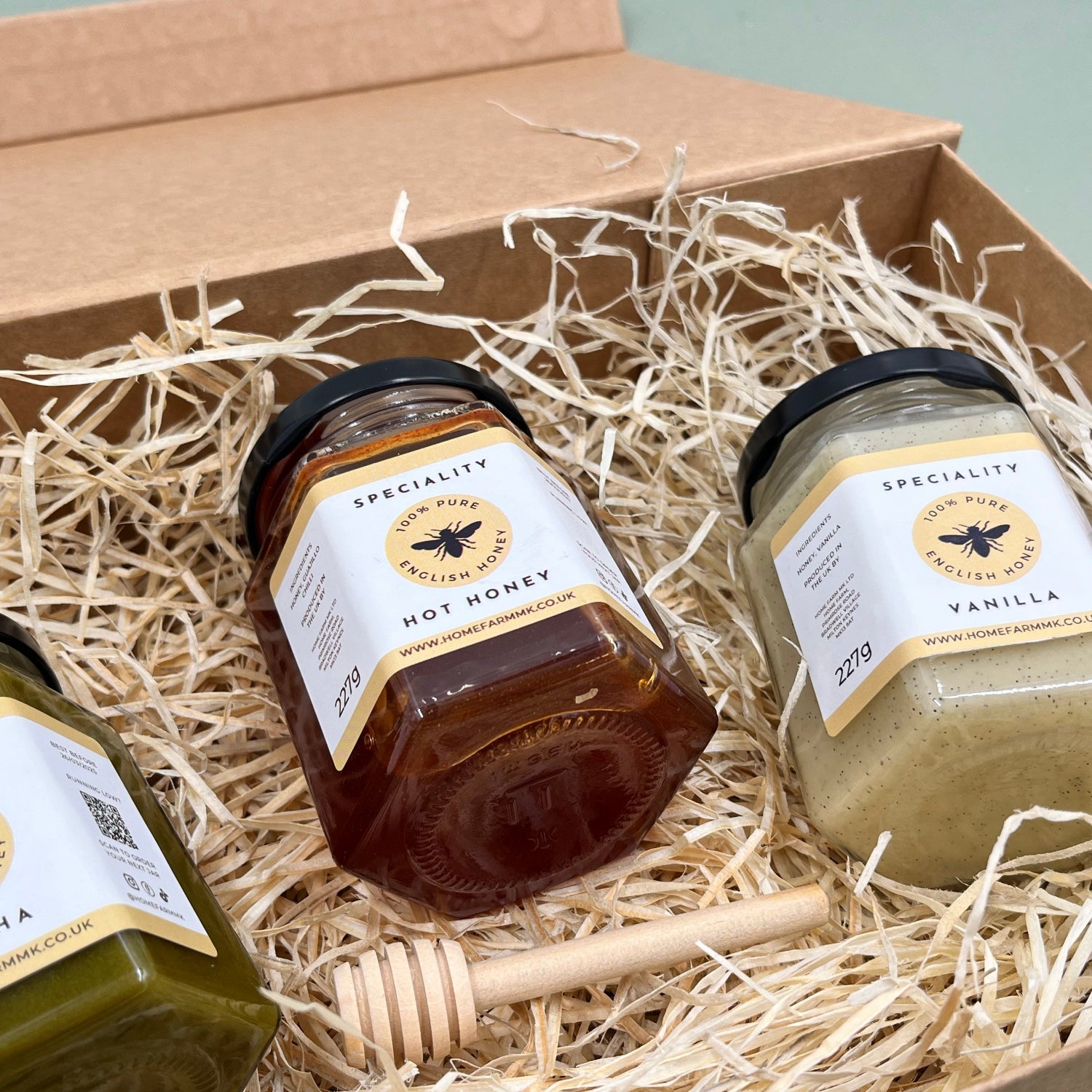 A jar of green matcha honey, a jar of red smoky hot honey, and a jar of white vanilla honey in a gift box, accompanied by a wooden honey dipper
