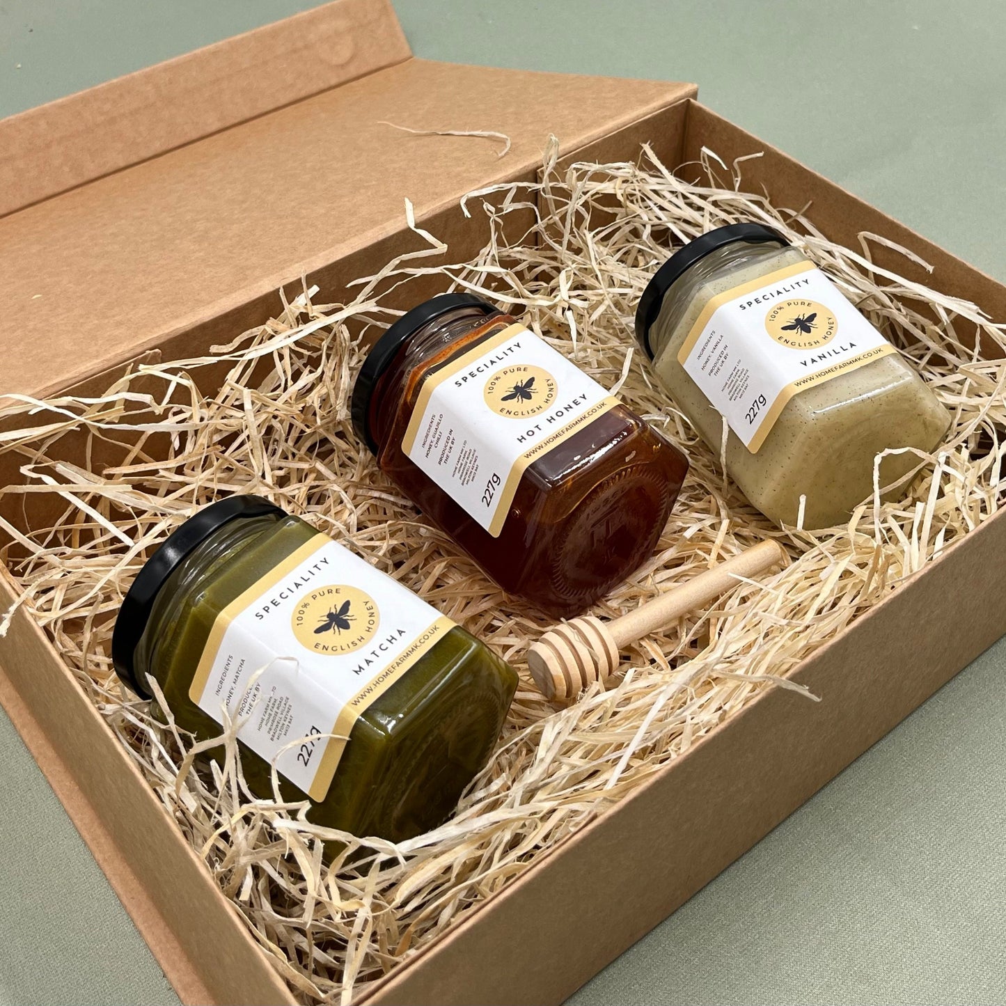 A jar of matcha honey, a jar of hot honey and a jar of vanilla honey with a wooden honey dipper in a kraft brown gift box
