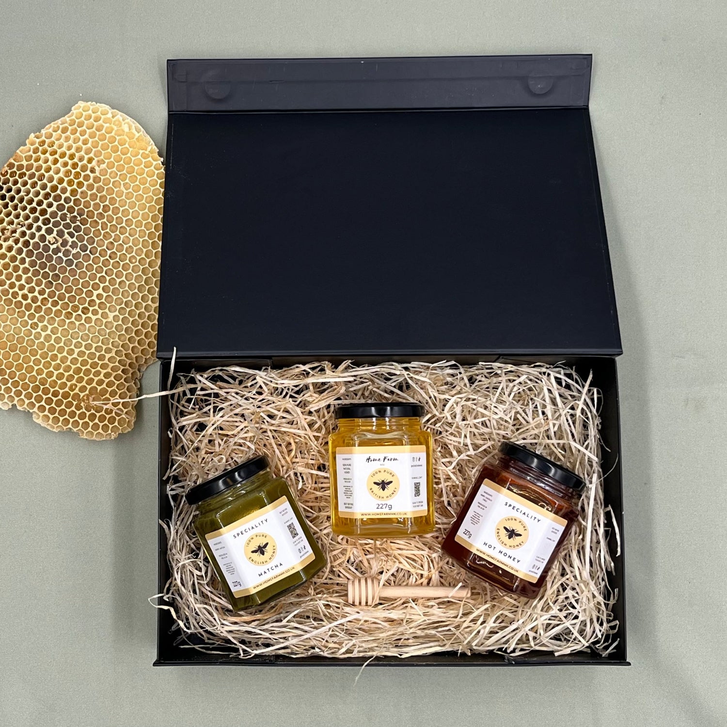 A jar of green matcha honey, a jar of yellow runny honey, and a jar of deep red smoky hot honey in a black gift-box, presented with a wooden honey dipper