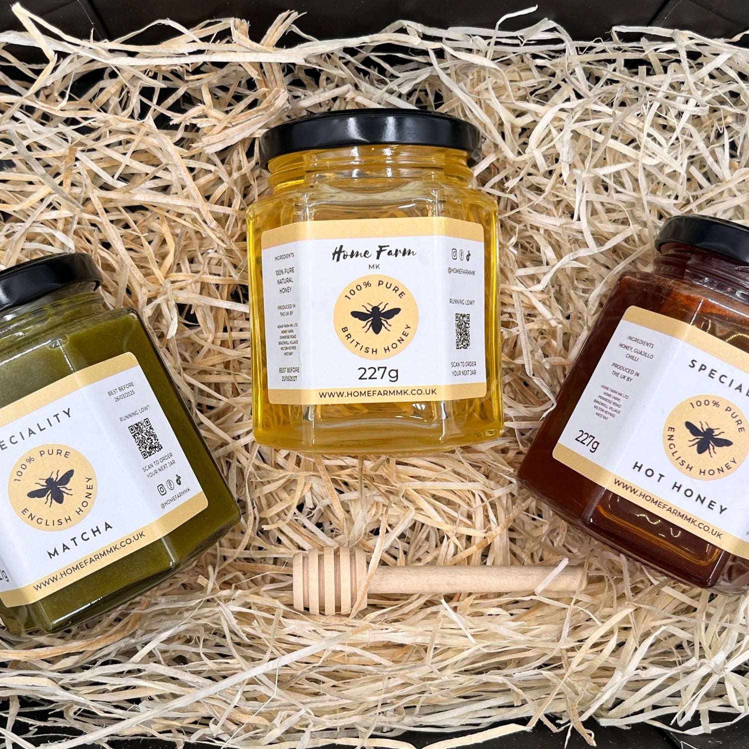 Three jars of honey and a wooden honey dipper on a bed of wood straw presented in a black gift box