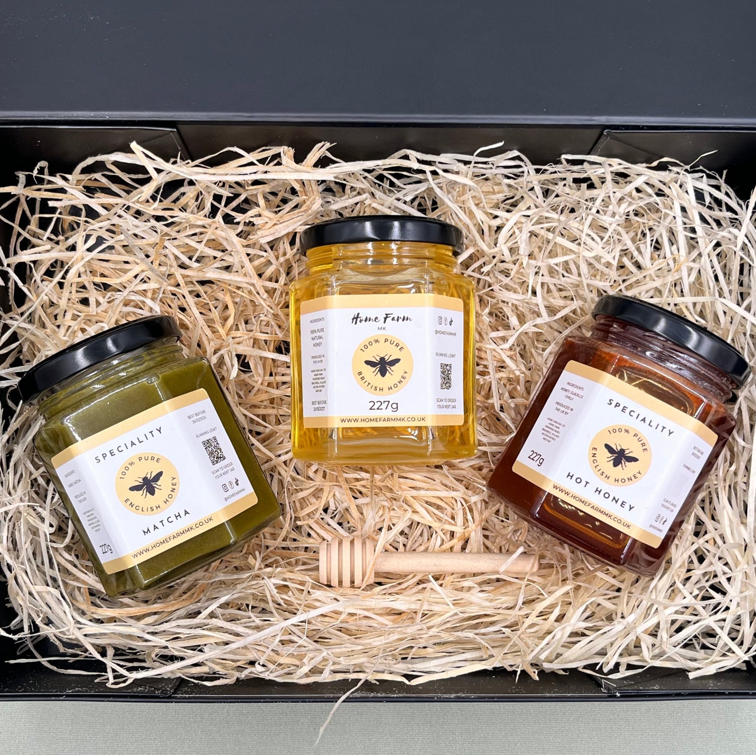 A jar of matcha honey, a jar of pure, English runny honey and a jar of hot honey with a wooden honey dipper in a black gift box.