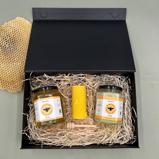 Gift set with two jars of honey and a yellow beeswax pillar candle and a wooden honey dipper in a black box.